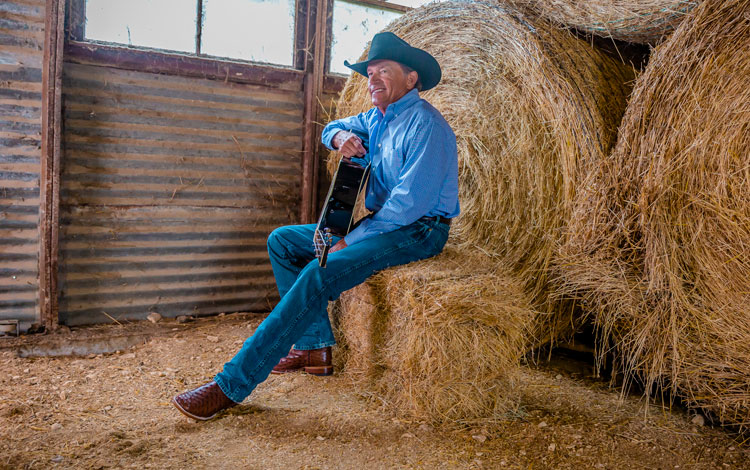 George Strait sitting on a square bale holding a guitar smiling.